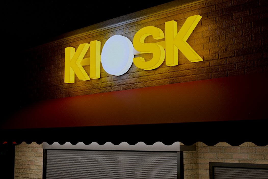 Kiosk sign with yellow letters on a brick wall at night