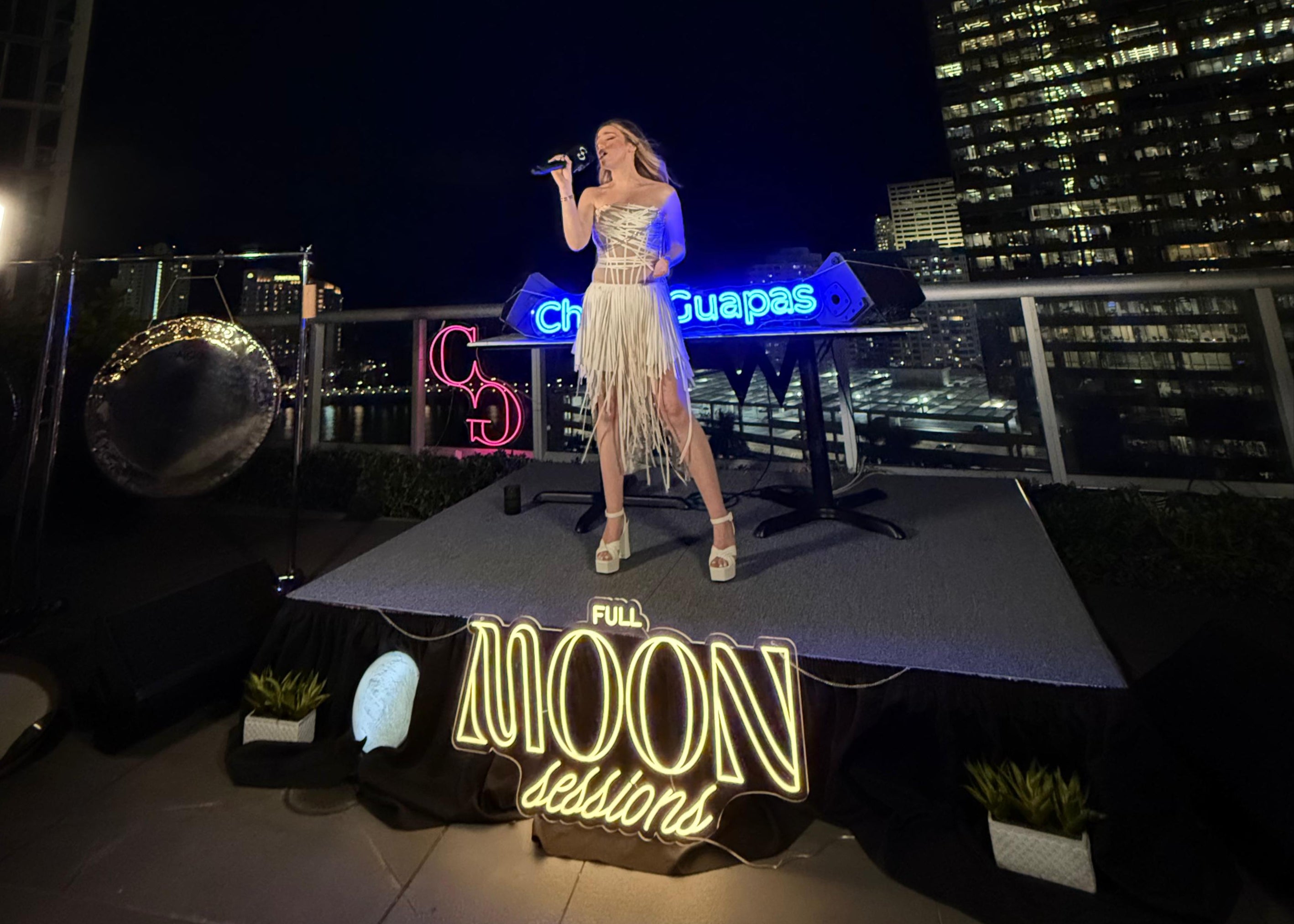 Woman on a stage with 'Full Moon Sessions' neon sign at a concert, cityscape in background
