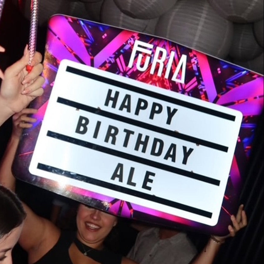 People celebrating a birthday with a 'Happy Birthday Ale' sign at a party.